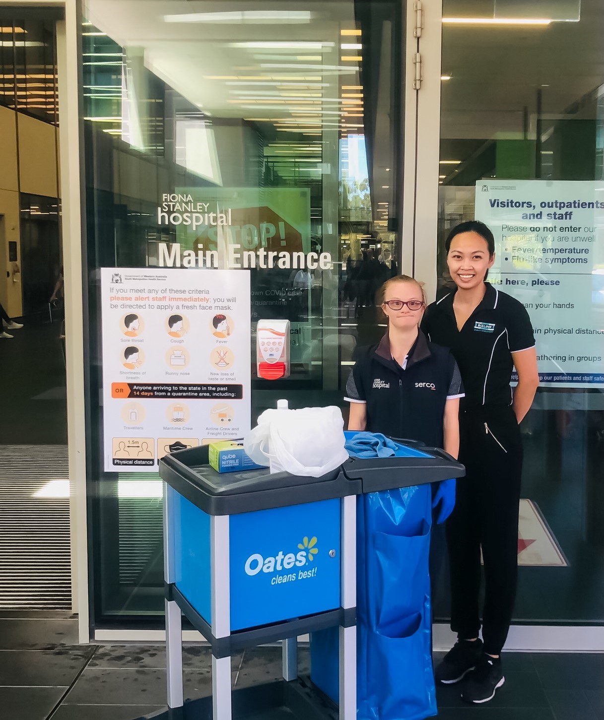 Sarah and her boss standing behind a cleaning trolley in front of a hospital