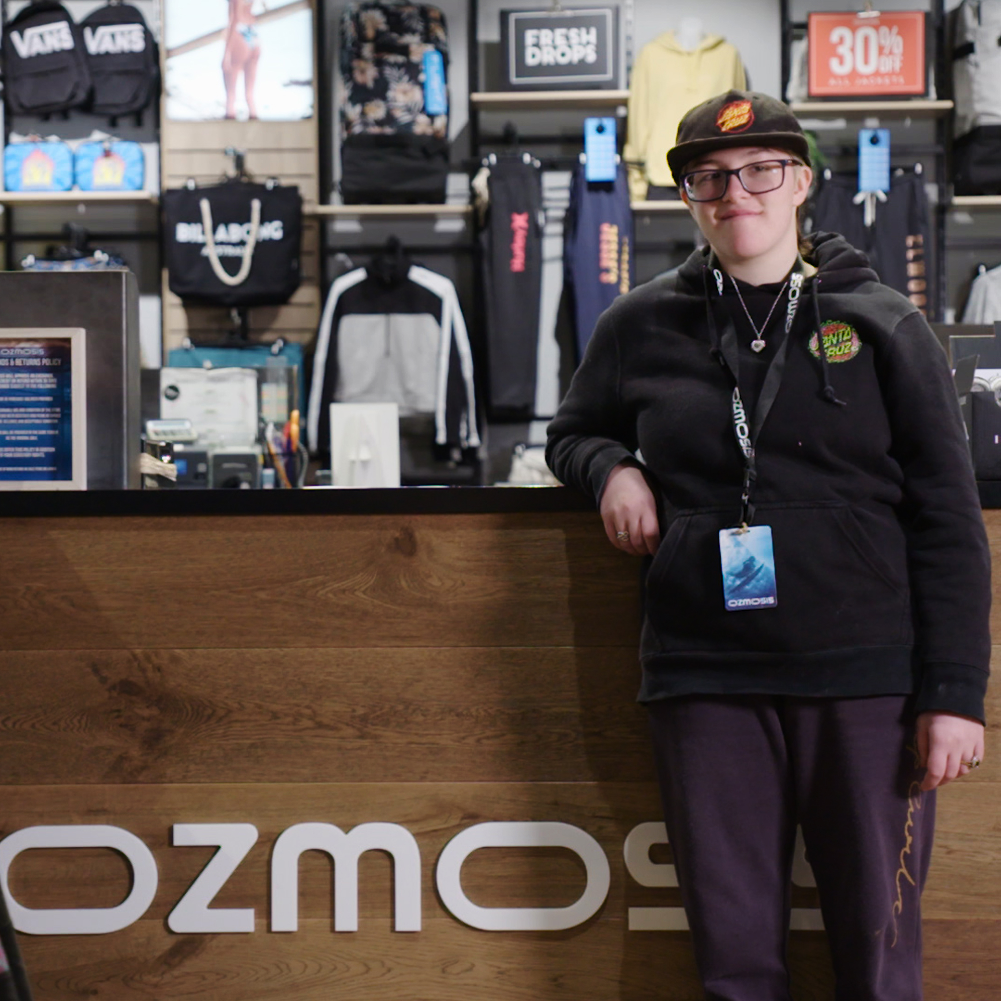 Danielle leaning on Ozmosis counter, wearing ozmosis clothes and has a lanyard around neck