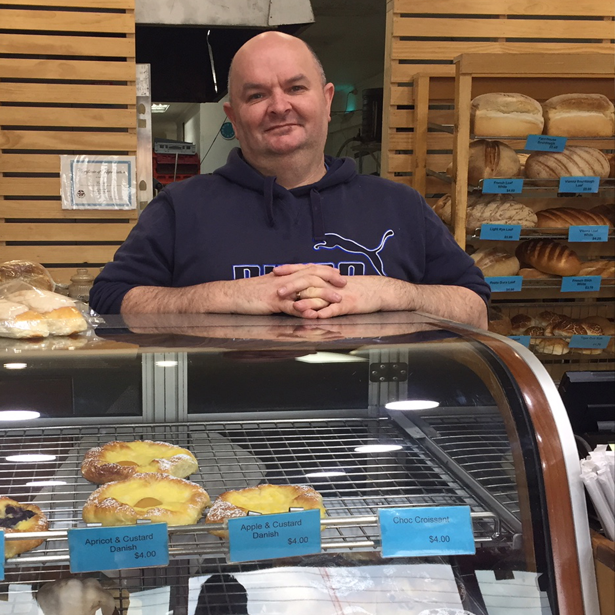 Dean with a small on his face and hand crossed standing behind a glass cabinet in a bakery with bread behind him on trays