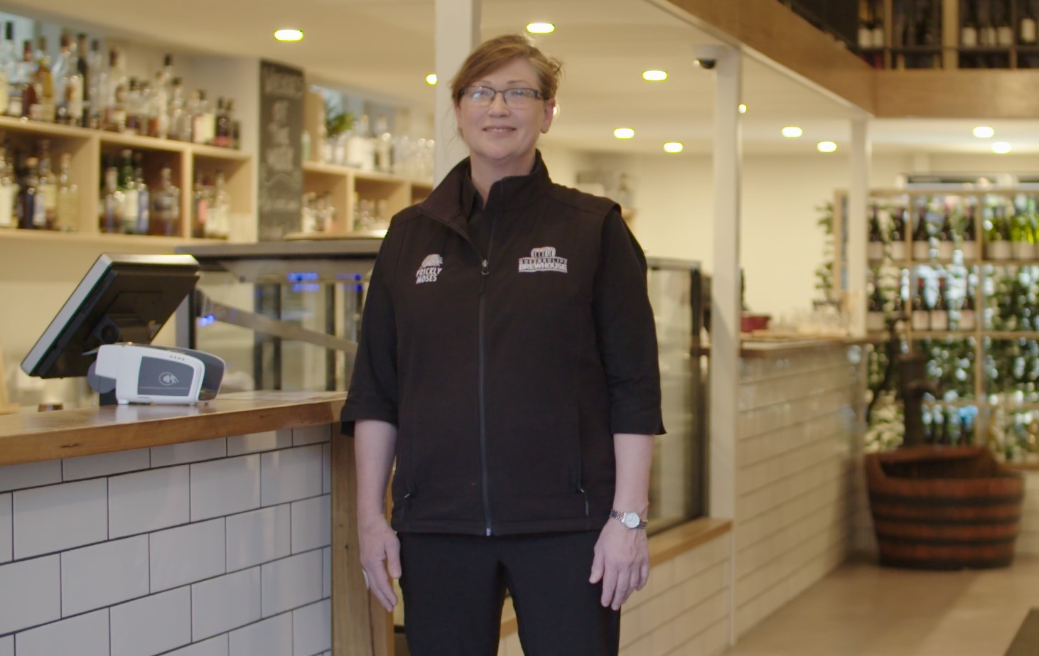 Deb smiling standing in a restaurant in front of serving area wearing black