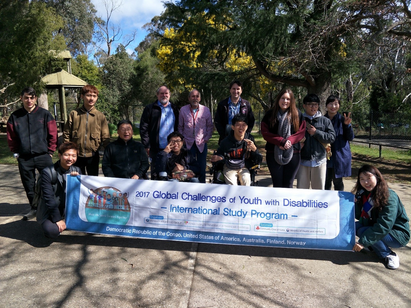A group students and teachers standing outside under trees with a banner in front saying global challenges of youth with disabilities