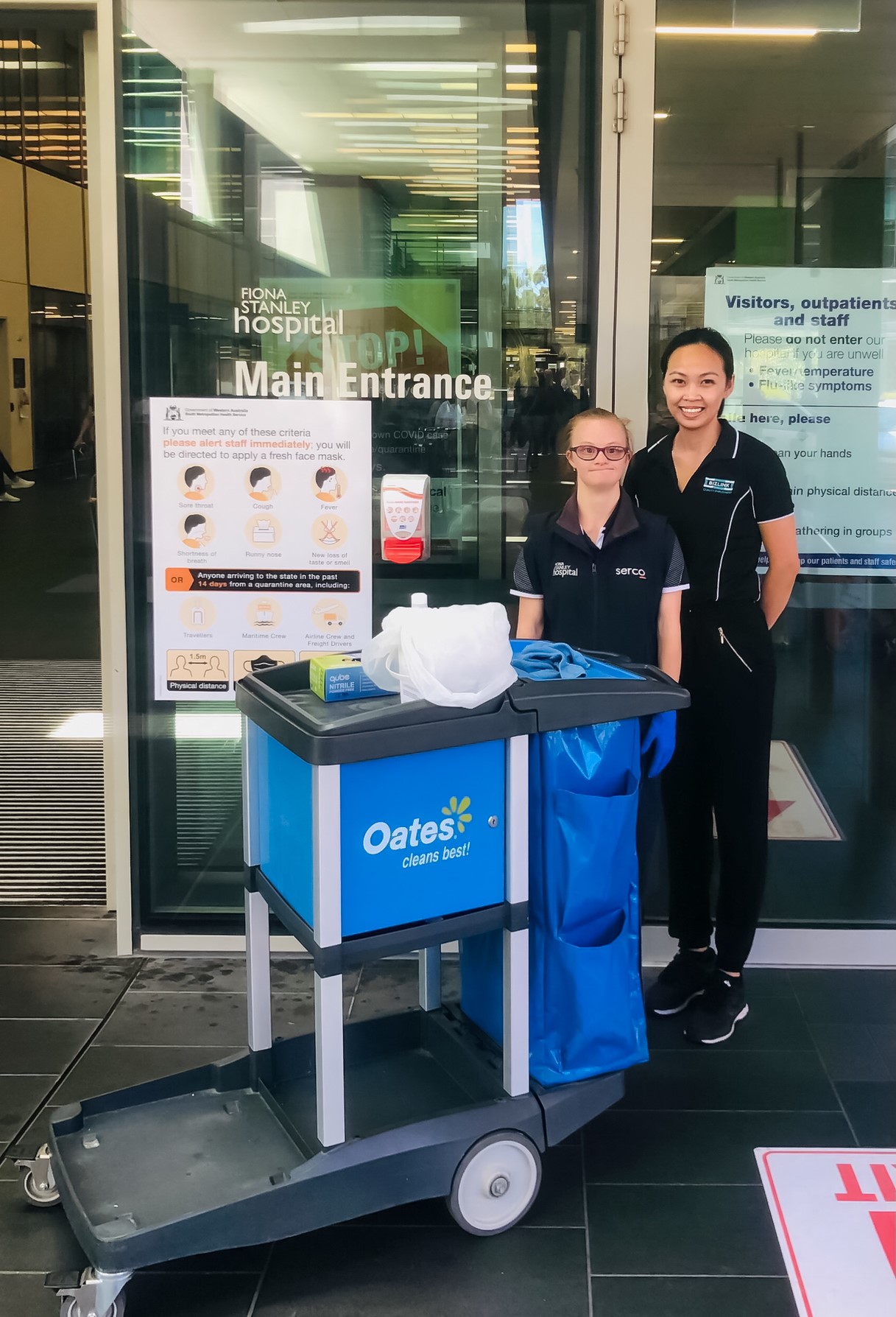 2 girls standing next to each other in front of a cleaning trolley and hospital entrance glass door