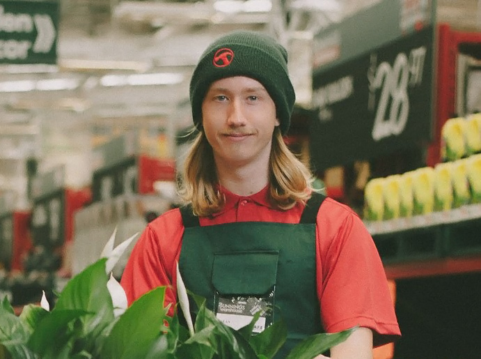 Sean standing in Bunnings with a Bunnings beanie  on a plant in front of him at bottom of image